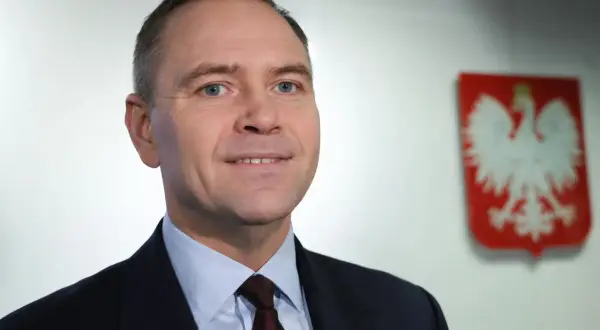 Polityk w garniturze z polskim godłem w tle.