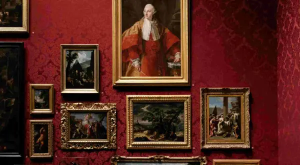 Galeria sztuki z obrazami w złotych ramach na czerwonej, wzorzystej ścianie. Widoczny portret mężczyzny w stroju z epoki, martwa natura z kwiatami i sceny rodzajowe. Różnorodność stylów ukazuje bogactwo nurtów w sztuce.