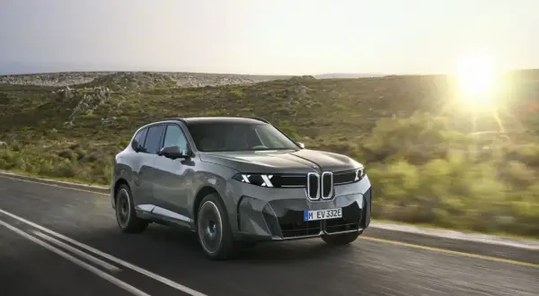El BMW iX5, un SUV eléctrico gris, avanza por una carretera rural al atardecer, con colinas verdes y el sol brillando.