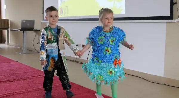 Chłopiec w eko stroju z opakowań po przekąskach i dziewczynka w sukience z kapsli i folii na pokazie mody.