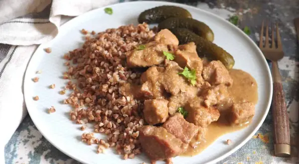 Gulasz z szynki wieprzowej w sosie własnym, podany z kaszą gryczaną i ogórkami kiszonymi. Pyszne, domowe danie.
