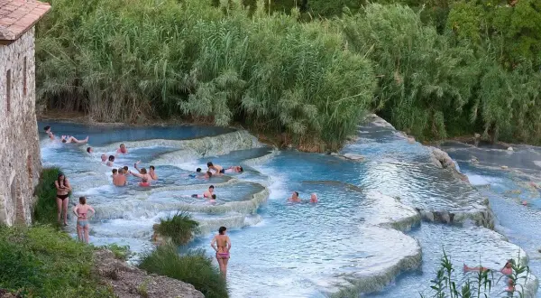 Persone si rilassano nelle cascate naturali dei aspio terme, con vegetazione lussureggiante e un antico edificio in pietra.