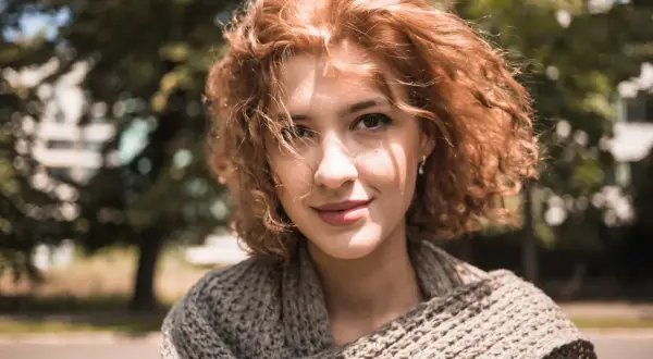 Uśmiechnięta rudowłosa dziewczyna w swetrze, idealna inspiracja, jak zrobić zdjęcie portretowe na tle drzew.