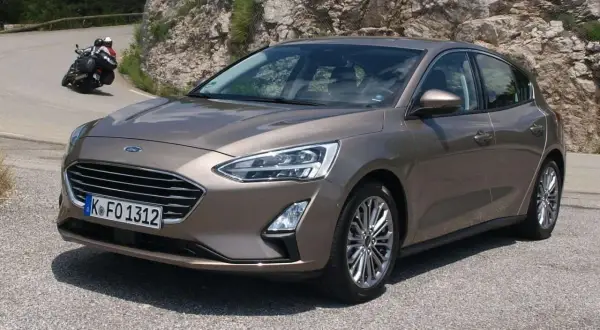 Brązowy Ford Focus 2017 na krętej drodze, z motocyklistą w tle.