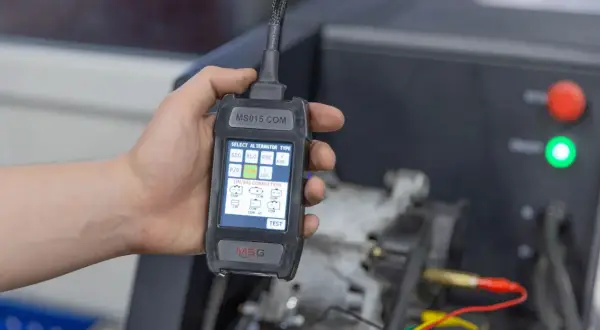 Dłoń trzyma tester diagnostyczny do alternator w samochodzie. Ekran pokazuje wybór typu alternatora i połączeń LIN/BSS.