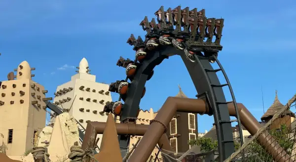 A roller coaster with brown seats and patterned spheres against a blue sky and themed buildings.