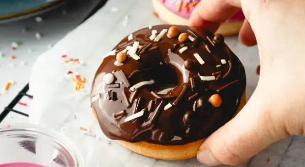 Leckeres Schoko-Donut-Rezept für den Backofen. Ein Donut mit Schokoglasur und Streuseln wird von einer Hand gehalten.