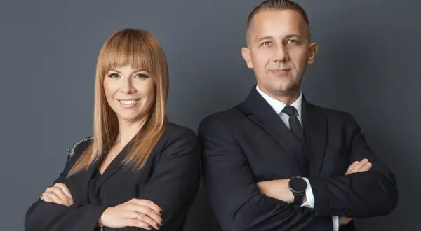 Dwie osoby w czarnych garniturach, kobieta i mężczyzna, stoją obok siebie ze skrzyżowanymi rękami.