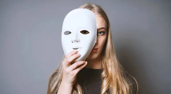 Una ragazza con lunghi capelli biondi tiene una maschera bianca davanti al viso. Forse un istrionico può amare, ma nasconde il suo vero volto.