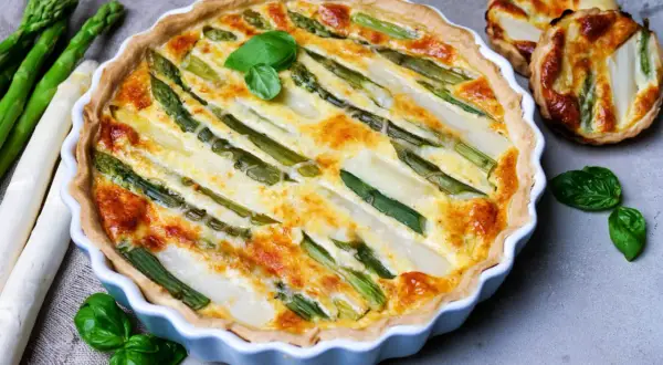 Herzhafte Spargel Quiche mit frischem Spargel und Kräutern, goldbraun gebacken. Daneben liegen roher Spargel und kleine Quiches.