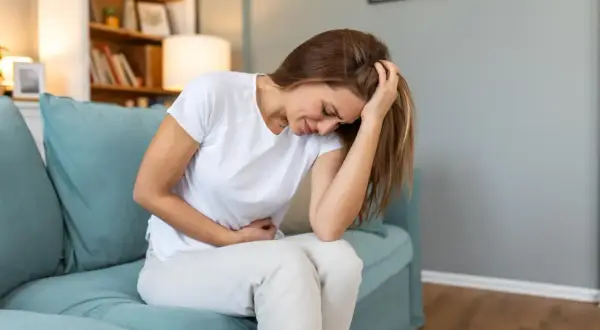 Femme assise sur un canapé bleu, se tenant le ventre et la tête, visiblement souffrante. Elle pourrait exprimer la douleur liée à l'endométriose profonde.