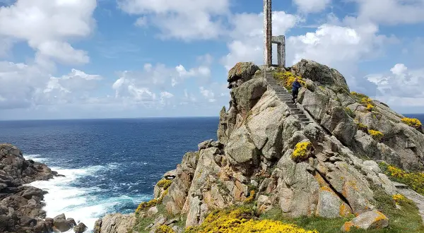 Escaleras de piedra ascienden por rocas cubiertas de flores amarillas hasta un monumento en el cabo Priorino. El mar azul y el cielo con nubes completan la escena.