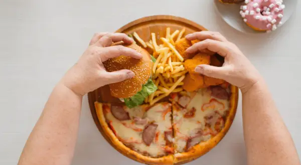 Mani che afferrano un hamburger, patatine, nuggets e pizza. Un vero **cheat day** con ciambelle in lontananza.