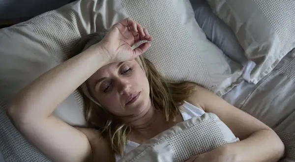 Mujer de unos 40 años sufre sudores nocturnos, con la mano en la frente, en la cama.