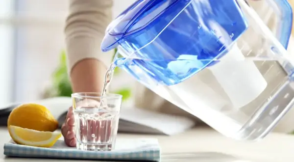 Une personne verse de l'eau filtrée d'une carafe bleue dans un verre. À côté, un citron coupé. L'eau filtrée est elle bonne pour la santé ?