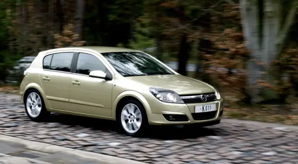 Złoty Opel Astra H 1.7 CDTI w ruchu na brukowanej drodze, otoczony rozmazanym tłem lasu.