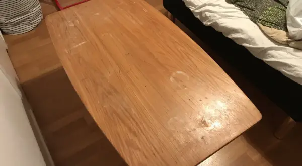 Ett slitet soffbord i trä med fläckar, redo att måla om. Bredvid syns en säng med vitt och grönt överkast.