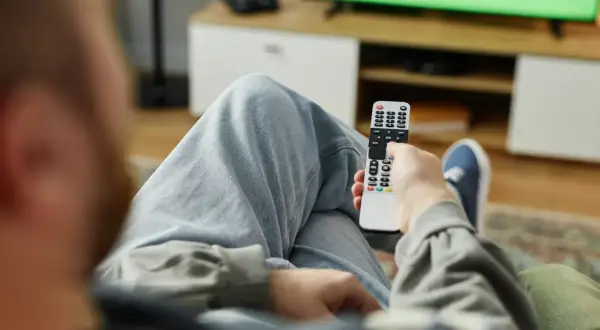 Mężczyzna z pilotem w ręku, zastanawia się, kto nie musi płacić abonamentu RTV, oglądając zielony ekran telewizora.