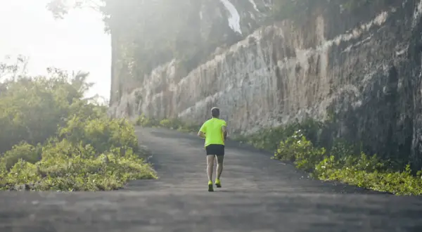 Un coureur en tenue fluo grimpe une route sinueuse, affrontant un dénivelé positif de 100 m, bordée de végétation luxuriante et d'une falaise imposante.