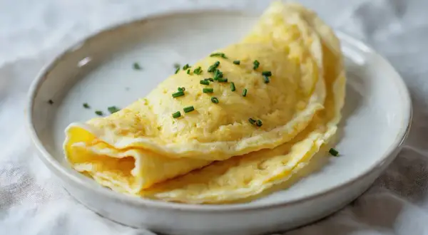 Delikatny omlet francuski, złożony na pół, posypany szczypiorkiem, na białym talerzu.
