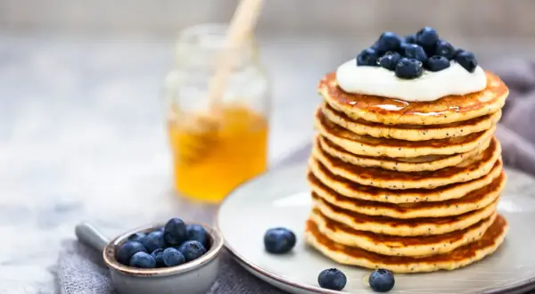 Pile de pancakes moelleux, garnis de crème et de myrtilles, avec un pot de miel en arrière-plan. Une recette pancake parfaite pour le petit-déjeuner.