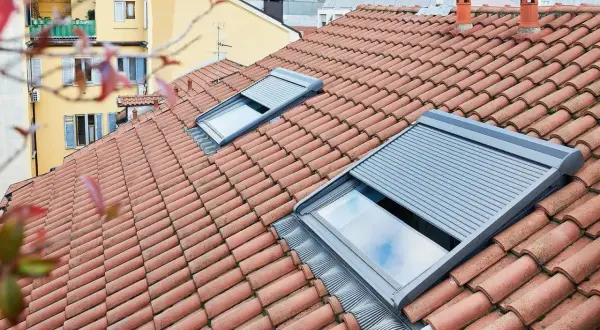Tegole rosse su un tetto con una pendenza tetto 30 gradi, due lucernari con tapparelle grigie parzialmente abbassate.