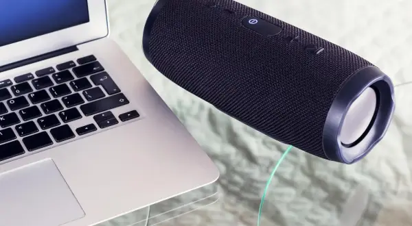 Un ordinateur portable et une enceinte Bluetooth noire sur une table en verre. Apprenez comment connecter une enceinte Bluetooth pour une expérience audio sans fil.