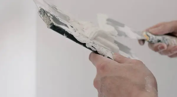 Händer som spacklar en vägg. Spacklet är vitt och klibbigt, och verktygen är täckta av det.