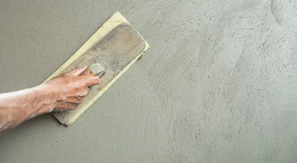 Mano che stende l'intonaco con una spatola, creando una finitura liscia. La rasatura 5 mm è quasi completa.
