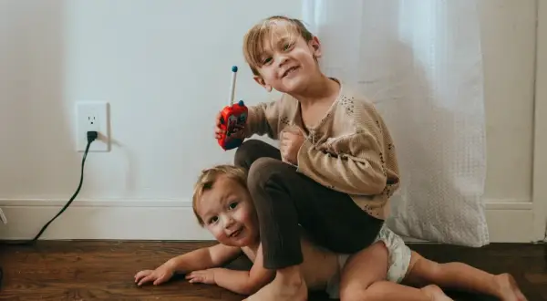 Twee broertjes spelen op de grond. De oudste zit op de rug van de jongste en houdt een speelgoedwalkietalkie vast. Dit is waarom maken broers en zussen ruzie.