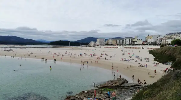 Gente disfruta de un día soleado en la playa de peizas, con aguas cristalinas y edificios al fondo.