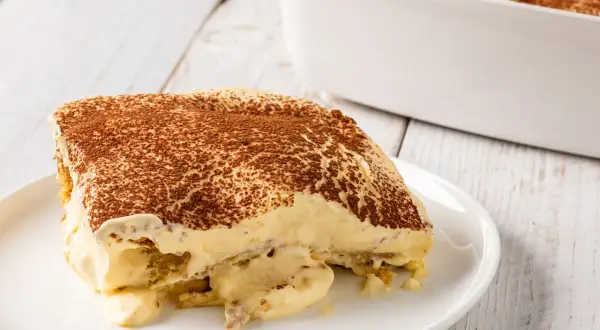 Una fetta di tiramisù, un classico dolce con mascarpone, cosparso di cacao amaro, su un piatto bianco.