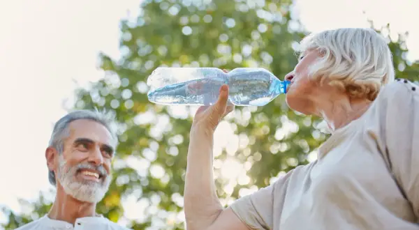 Un couple profite du soleil, la femme boit de l'eau pour une hydratation optimale, l'homme sourit.