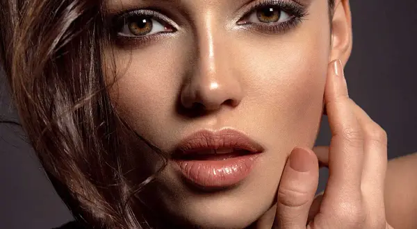 Primo piano di una donna con un sofisticato trucco occhi neri, labbra lucide e capelli castani mossi.