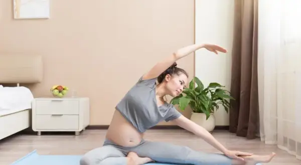 Ciężarna kobieta wykonuje ćwiczenie rozciągające statyczne na macie w sypialni.