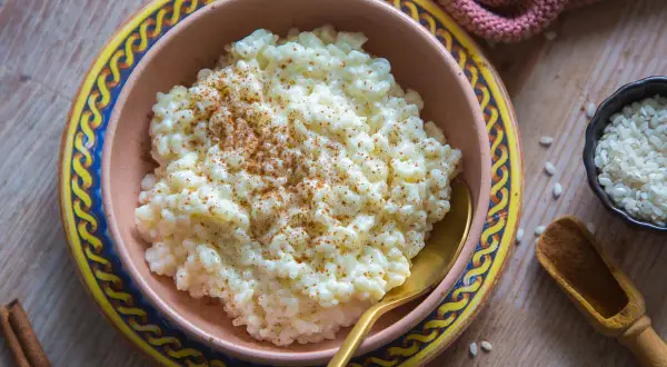 Omas Milchreis mit Butter, bestreut mit Zimt, in einer Schüssel auf einem Holztisch. Daneben eine kleine Schale Reis und eine Zimtstange.