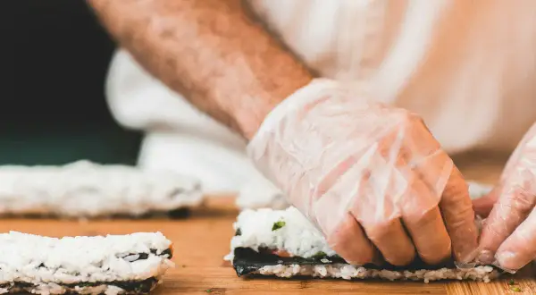 Hände mit Handschuhen rollen Sushi. Reis, Nori und Gemüse sind die Zutaten für sushi selber machen.