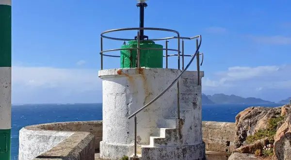 Faro blanco con base verde y escaleras externas, junto al mar azul. El **monte Ferro** se divisa al fondo.