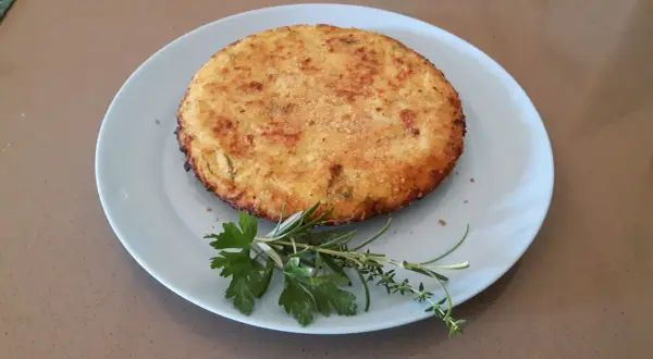 Frittata di cous cous dorata e croccante, guarnita con erbe aromatiche fresche su un piatto azzurro.