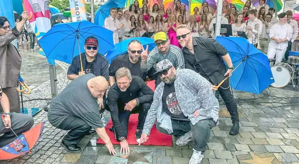 Grupa mężczyzn, prawdopodobnie zespół, odsłania gwiazdę z napisem "PIERSI BAŁKANICA" na chodniku. W tle kolorowe parasole i publiczność.