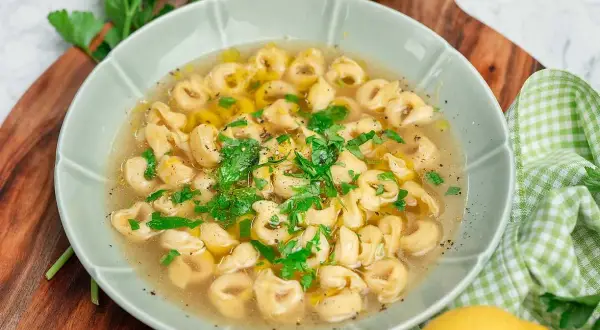 En skål med tortellini in brodo, garnerad med färsk persilja och en citronskiva.