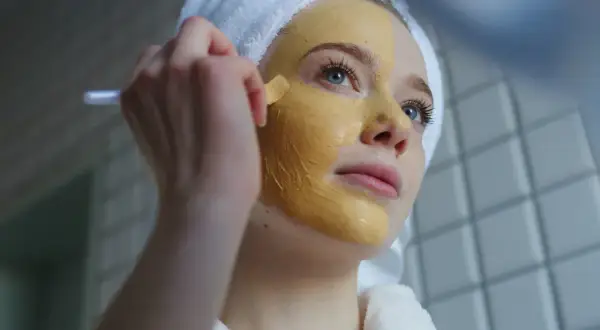 Junge Frau mit Handtuch auf dem Kopf trägt eine Kurkuma-Maske auf. Vorher-Nachher-Effekt für strahlende Haut.