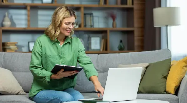 Kobieta na kanapie z laptopem i notatkami uczy się, jak doładować prąd przez internet.