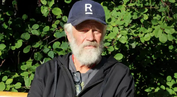 En äldre man med vitt skägg och keps med ett R på sitter utomhus. Han är en del av Ronny Eriksson familj.