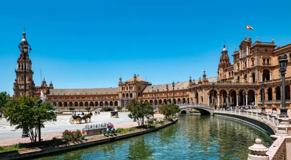 Vacker vy över Plaza de España i Sevilla, Spanien. En hästdragen vagn glider fram längs kanalen under en klarblå himmel.