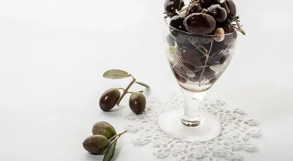 Un bicchiere pieno di olive nere, con rametti e foglie, accanto a olive fresche su un pizzo. Le olive delle Marche, un sapore autentico.