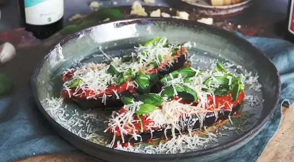 Två halvor av aubergine i ugn, toppade med tomatsås, basilika och riven ost. Serveras på en rustik tallrik.