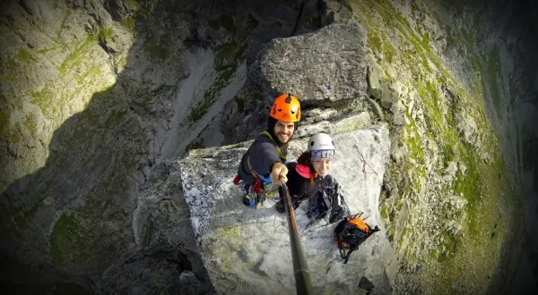Uśmiechnięci alpiniści na skalnej półce, gotowi na wejście na mnicha. Widok z góry na stromą ścianę.