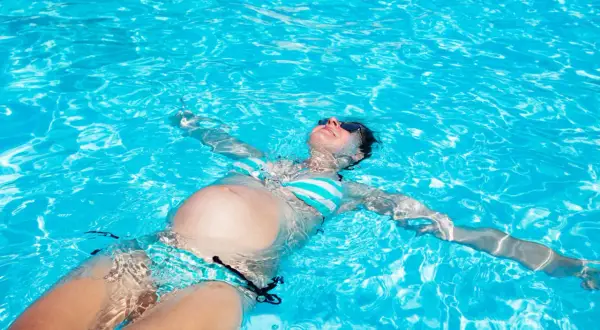 Eine schwangere Frau entspannt sich beim **baden in der Schwangerschaft** auf dem Rücken im türkisfarbenen Poolwasser.