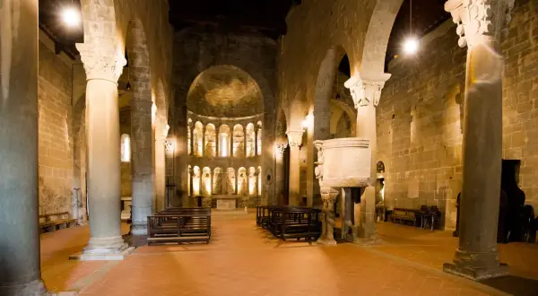 Interno della pieve di Gropina, con colonne massicce, archi in pietra, un pulpito scolpito e l'abside illuminata.